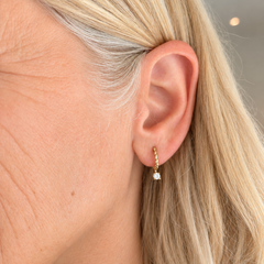 A close-up side profile of a woman with blonde hair wearing a delicate gold huggie hoop earring with a small diamond dangling from the earring
