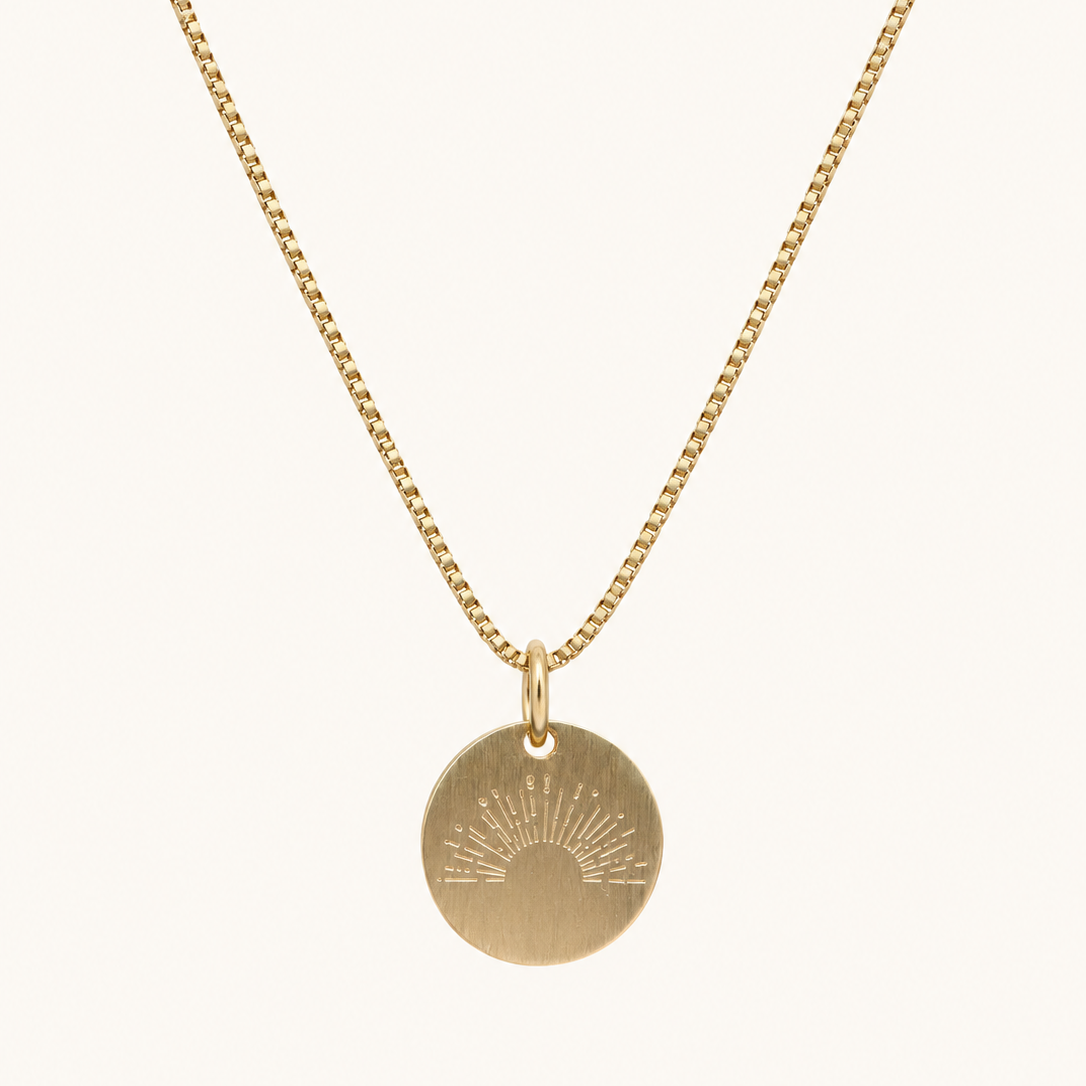 A gold chain necklace with a round disc pendant featuring an engraved sunburst design, shown against a plain off-white background.