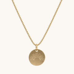 A gold chain necklace with a round disc pendant featuring an engraved sunburst design, shown against a plain off-white background.