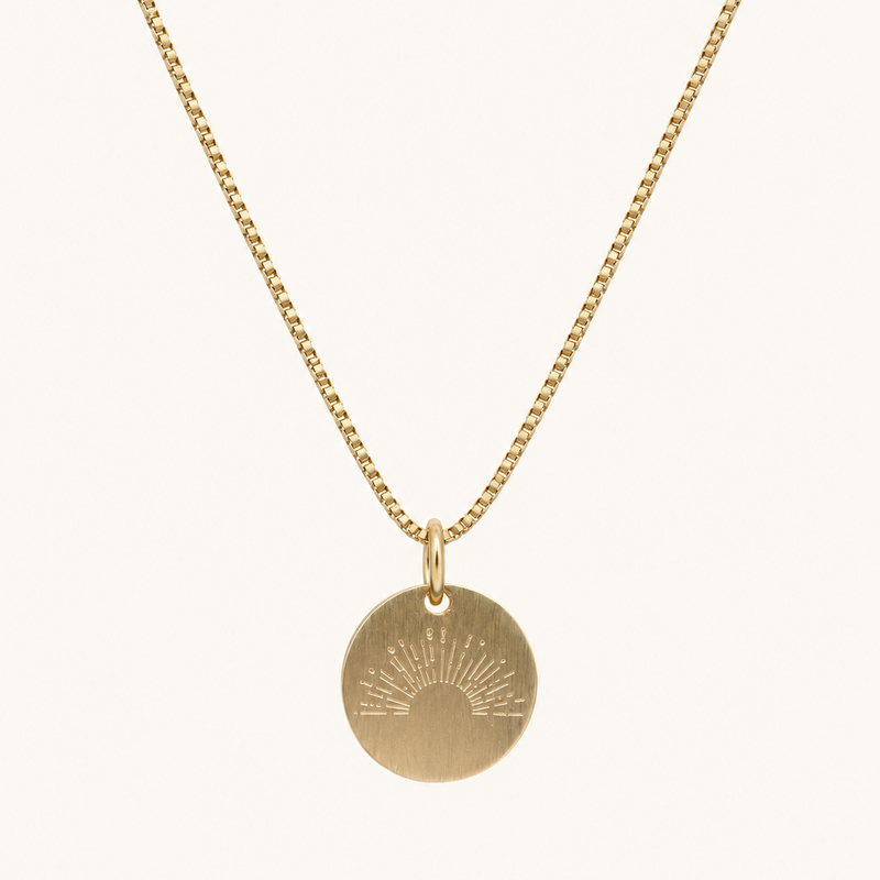 A gold chain necklace with a round disc pendant featuring an engraved sunburst design, shown against a plain off-white background.