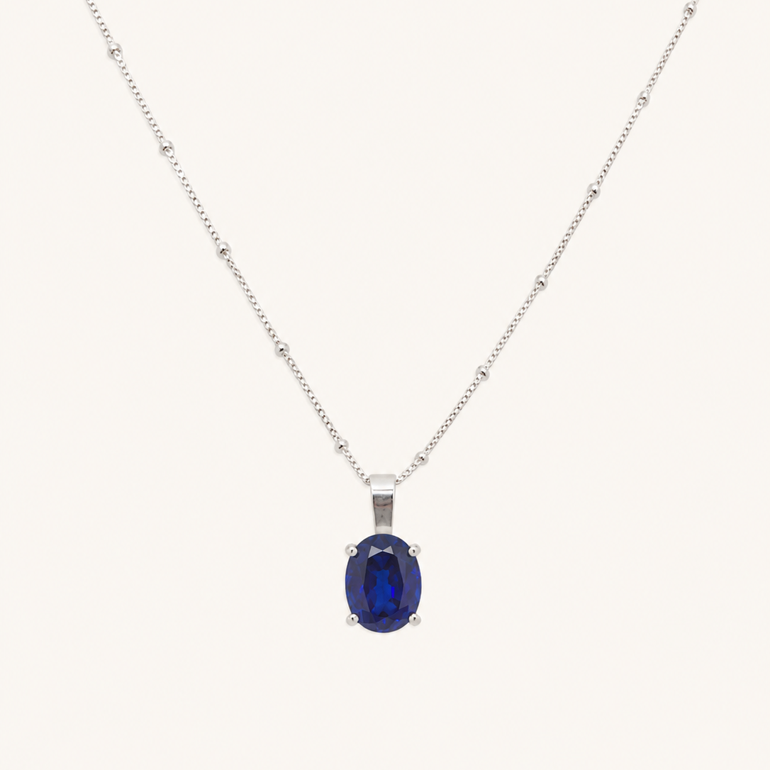A silver chain necklace with a central oval dark blue gemstone pendant, displayed against a solid off-white background.