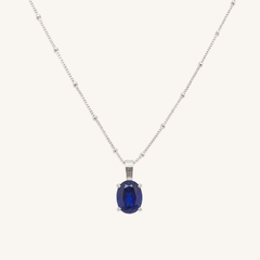 A silver chain necklace with a central oval dark blue gemstone pendant, displayed against a solid off-white background.