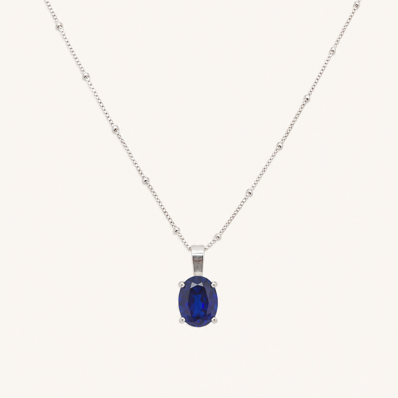 A silver chain necklace with a central oval dark blue gemstone pendant, displayed against a solid off-white background.