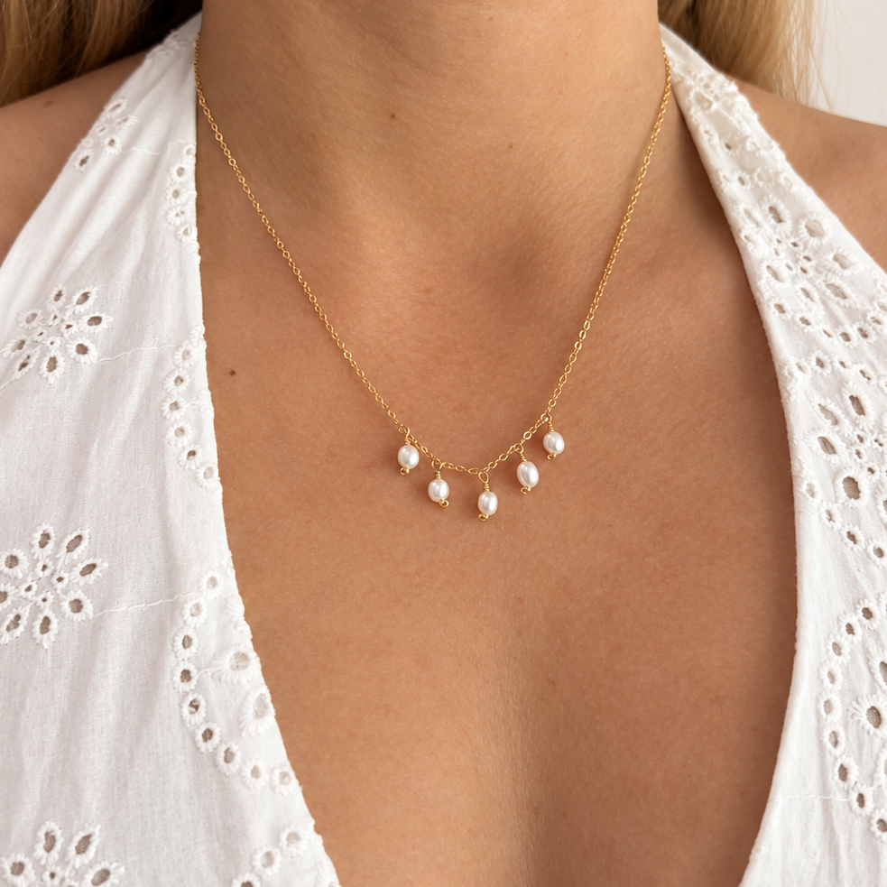 A close-up of a model wearing a delicate gold chain necklace with five small white pearls, styled with a white eyelet halter top.