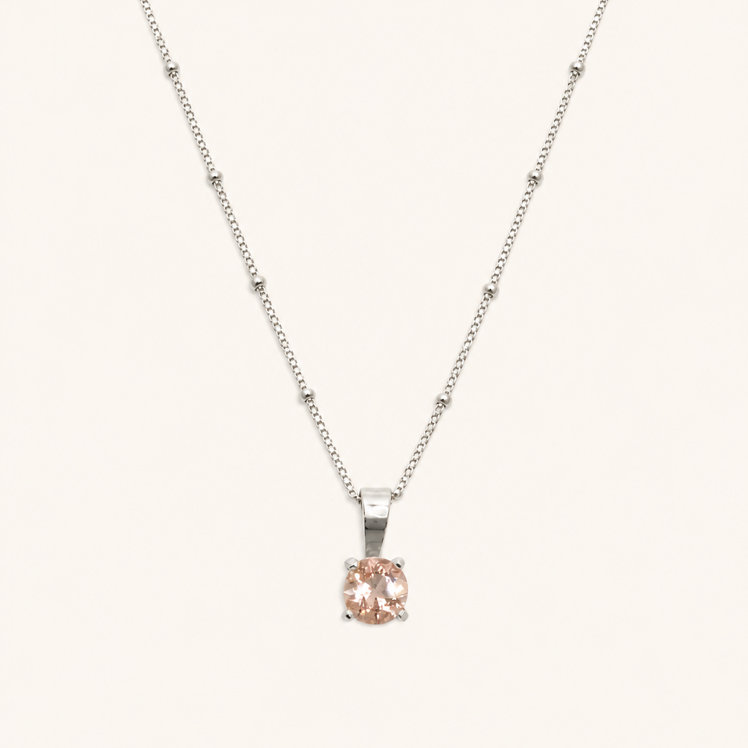 A delicate silver necklace with a small, round-cut pale pink gemstone pendant, displayed against a solid off-white background.