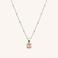 A delicate silver necklace with a small, round-cut pale pink gemstone pendant, displayed against a solid off-white background.