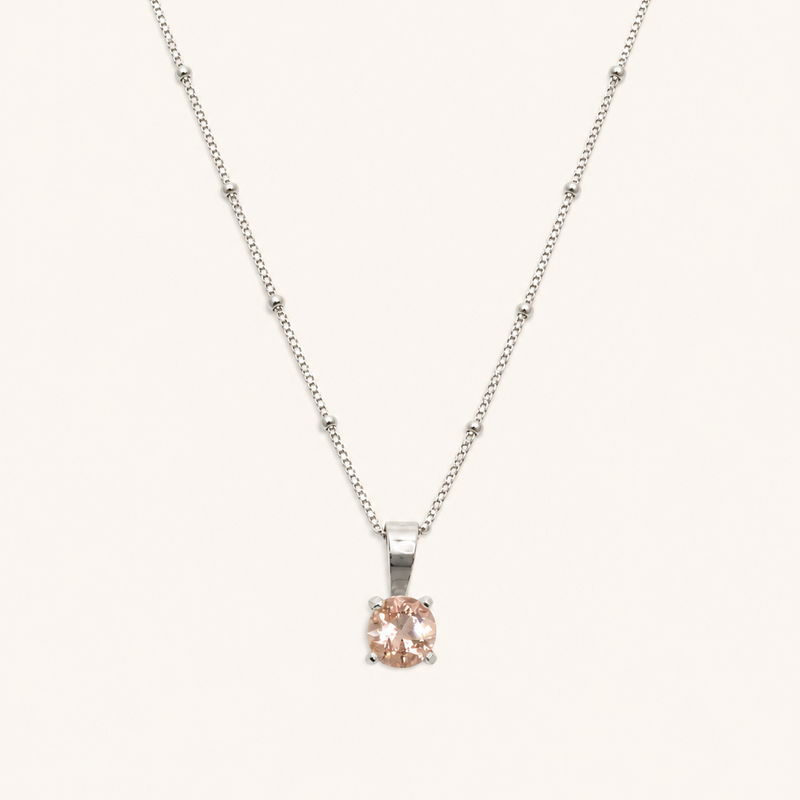 A delicate silver necklace with a small, round-cut pale pink gemstone pendant, displayed against a solid off-white background.