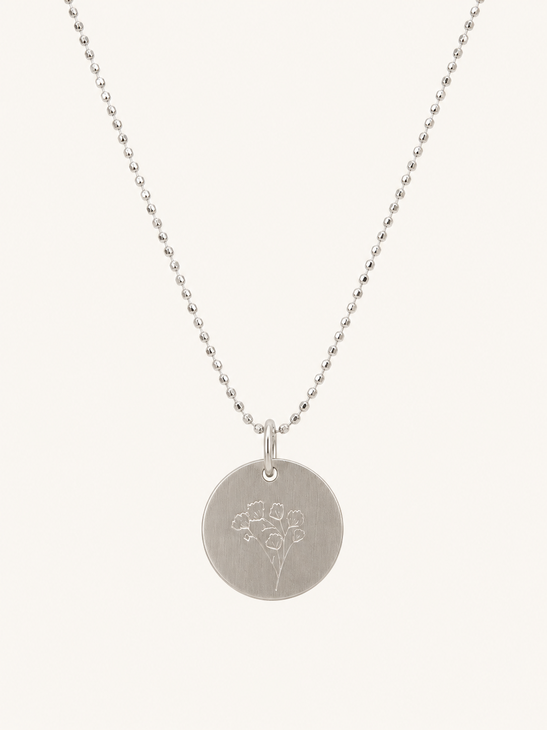 A silver disc pendant necklace with a delicate floral engraving, suspended from a fine silver ball chain against a light cream background.