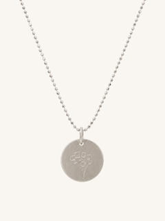 A silver disc pendant necklace with a delicate floral engraving, suspended from a fine silver ball chain against a light cream background.