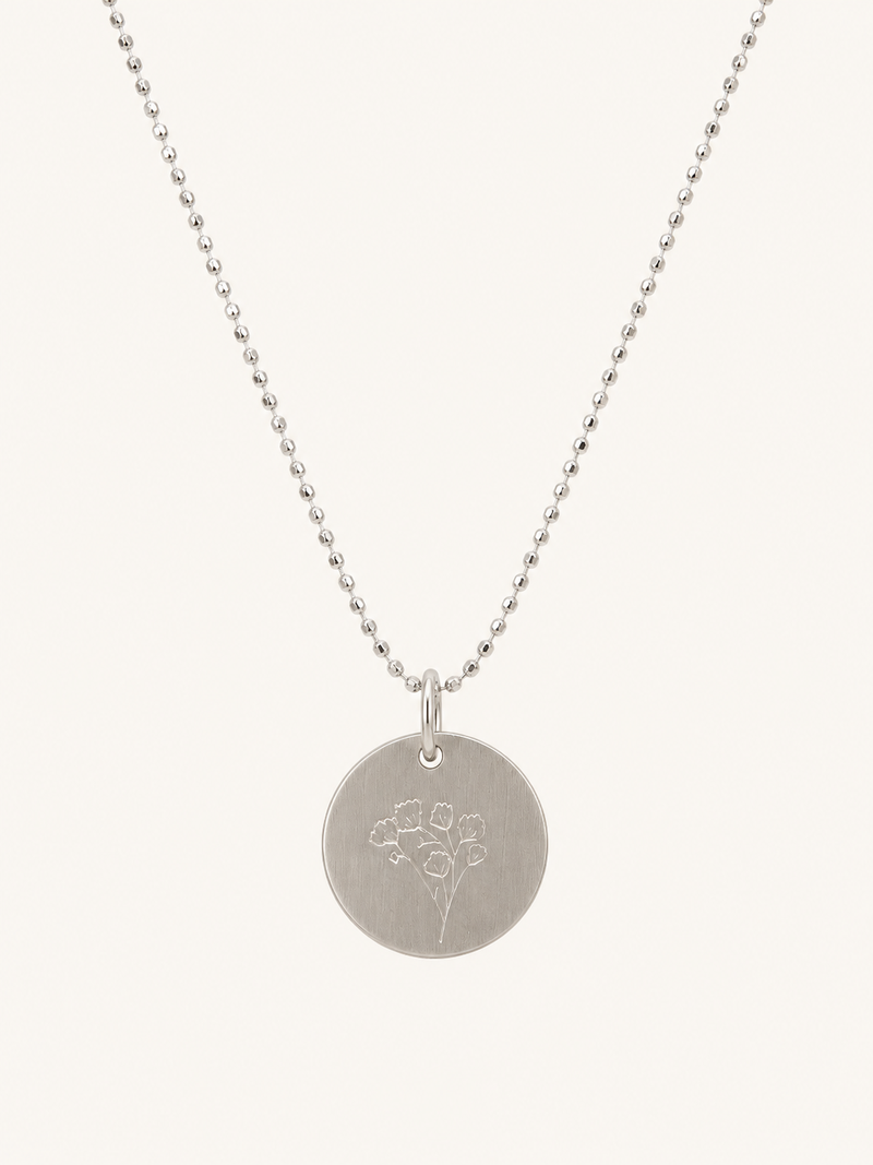 A silver disc pendant necklace with a delicate floral engraving, suspended from a fine silver ball chain against a light cream background.