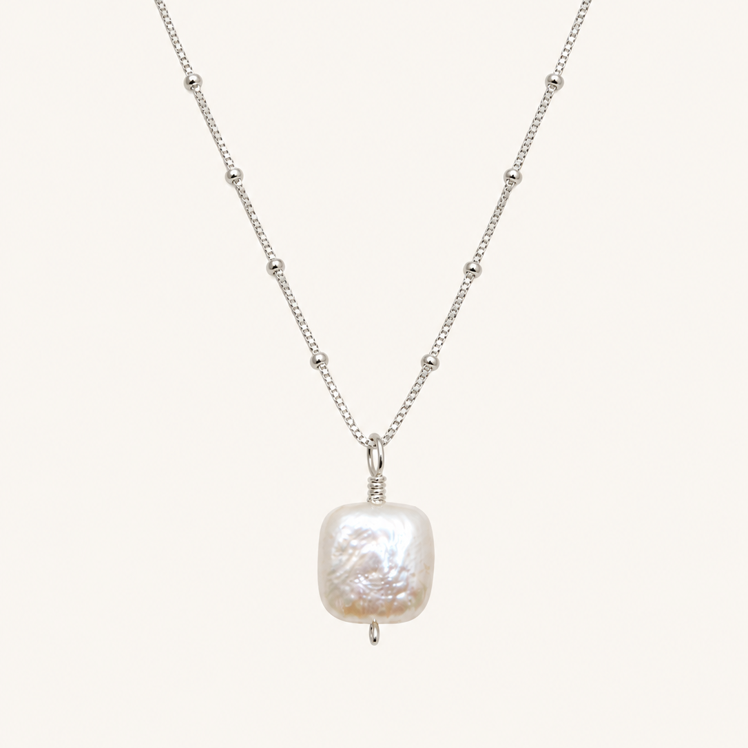 A silver beaded chain necklace featuring a large, irregularly shaped white baroque pearl pendant, displayed against a solid off-white background.